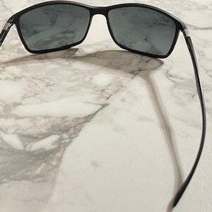 May include: Black sunglasses with rectangular lenses and a thin, black frame. The sunglasses are positioned on a light-colored, marbled surface, casting a shadow. The arms of the sunglasses extend outward, creating a symmetrical appearance.