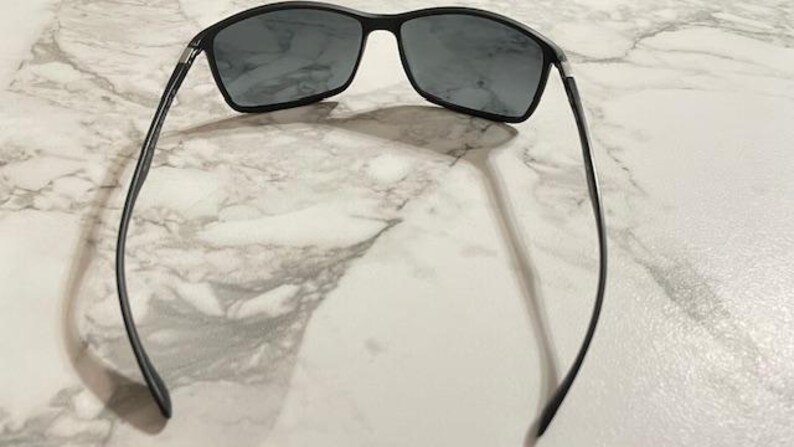 May include: Black sunglasses with rectangular lenses and a thin, black frame. The sunglasses are positioned on a light-colored, marbled surface, casting a shadow. The arms of the sunglasses extend outward, creating a symmetrical appearance.