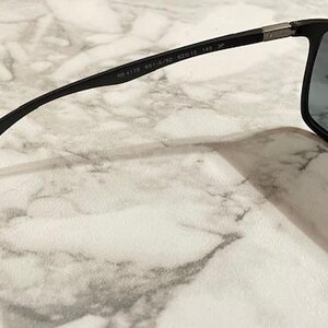 May include: Black sunglasses with rectangular lenses and a sleek, modern design. The frame is black with a subtle silver accent near the hinges. The lenses are a dark gray color, providing shade and style.