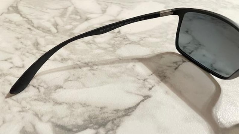 May include: Black sunglasses with rectangular lenses and a sleek, modern design. The frame is black with a subtle silver accent near the hinges. The lenses are a dark gray color, providing shade and style.