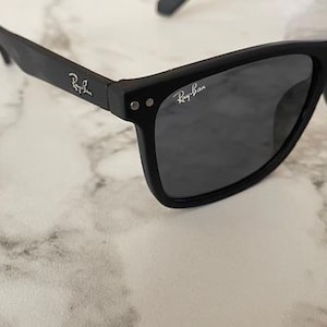 Pu&ograve; includere: Occhiali da sole neri con lenti grigio scuro. Il logo Ray-Ban &egrave; visibile sulle aste e sulla lente. Gli occhiali da sole sono appoggiati su una superficie di marmo bianco e grigio.