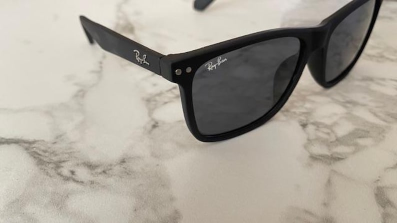 Pu&ograve; includere: Occhiali da sole neri con lenti grigio scuro. Il logo Ray-Ban &egrave; visibile sulle aste e sulla lente. Gli occhiali da sole sono appoggiati su una superficie di marmo bianco e grigio.
