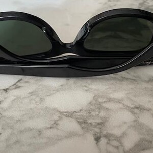 K&ouml;nnte beinhalten: Schwarze Sonnenbrille mit dunkelgr&uuml;nen Gl&auml;sern. Die Sonnenbrille ist zusammengeklappt und zeigt den Innenrahmen und das Ray-Ban-Logo am B&uuml;gel. Die Brille liegt auf einer marmorierten Oberfl&auml;che.