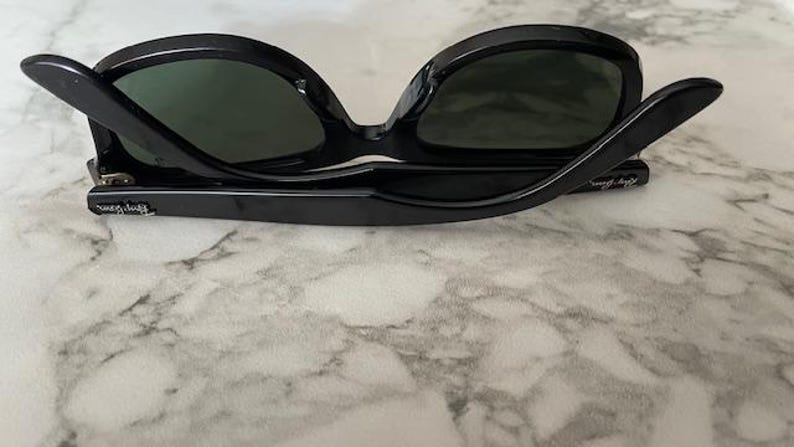 K&ouml;nnte beinhalten: Schwarze Sonnenbrille mit dunkelgr&uuml;nen Gl&auml;sern. Die Sonnenbrille ist zusammengeklappt und zeigt den Innenrahmen und das Ray-Ban-Logo am B&uuml;gel. Die Brille liegt auf einer marmorierten Oberfl&auml;che.