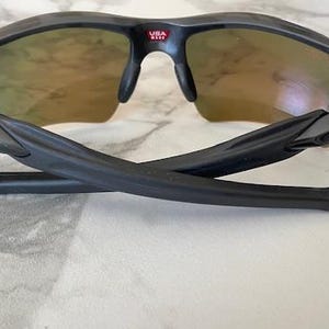 Peut inclure: Lunettes de soleil de sport noires avec un design semi-cercl&eacute;. Les verres ont une teinte d&eacute;grad&eacute;e, passant d'une teinte dor&eacute;e en haut &agrave; une teinte plus claire. La monture est noire avec le texte "USA" en rouge. Les branches sont pli&eacute;es.
