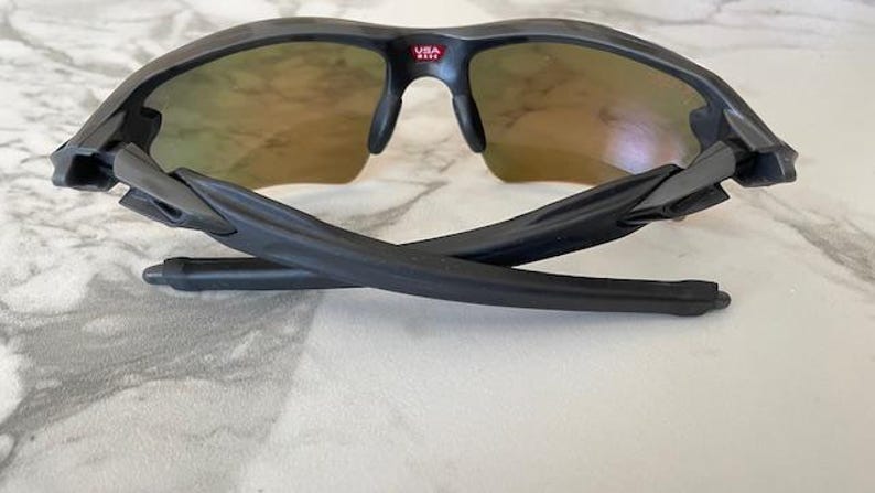 Peut inclure: Lunettes de soleil de sport noires avec un design semi-cercl&eacute;. Les verres ont une teinte d&eacute;grad&eacute;e, passant d'une teinte dor&eacute;e en haut &agrave; une teinte plus claire. La monture est noire avec le texte "USA" en rouge. Les branches sont pli&eacute;es.