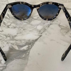 Puede incluir: Gafas de sol redondas con montura con estampado de carey y lentes azules. Las gafas de sol est&aacute;n colocadas sobre un fondo de m&aacute;rmol. Las patillas de las gafas de sol son curvas y se extienden hacia afuera.