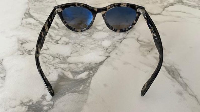 Puede incluir: Gafas de sol redondas con montura con estampado de carey y lentes azules. Las gafas de sol est&aacute;n colocadas sobre un fondo de m&aacute;rmol. Las patillas de las gafas de sol son curvas y se extienden hacia afuera.