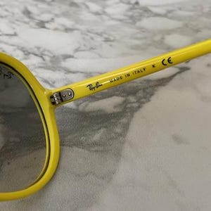 May include: Close-up of yellow sunglasses with gray lenses. The sunglasses have the Ray-Ban logo and the text "MADE IN ITALY" printed on the arm. The frame is a bright yellow color.