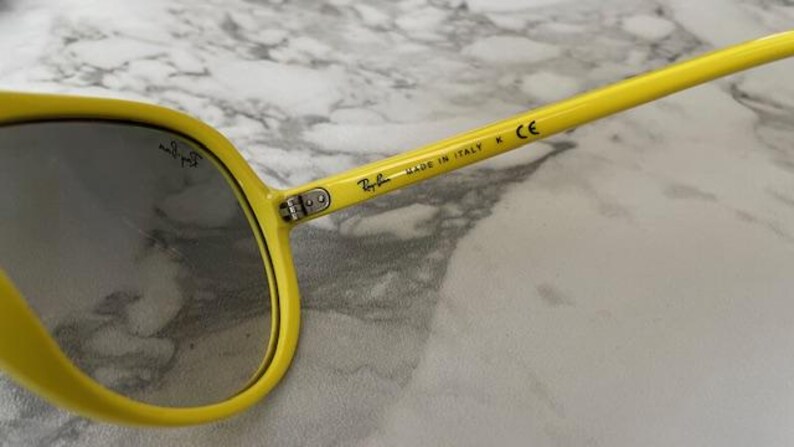 May include: Close-up of yellow sunglasses with gray lenses. The sunglasses have the Ray-Ban logo and the text "MADE IN ITALY" printed on the arm. The frame is a bright yellow color.