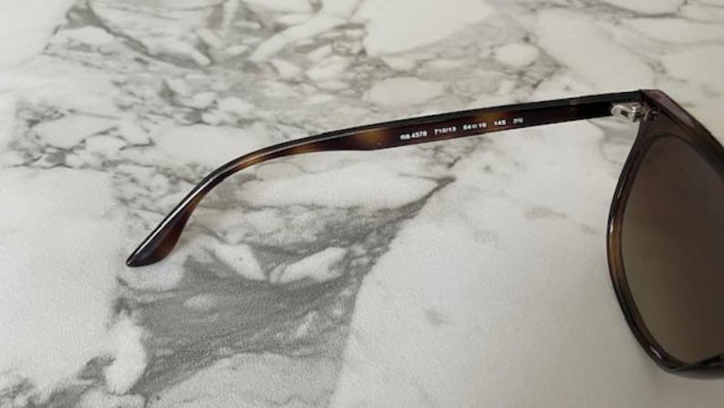 May include: Close-up of sunglasses with a brown tortoiseshell frame and gradient lenses. The arm of the glasses has the model number RB 4378, along with other details. The glasses are set against a marble background.
