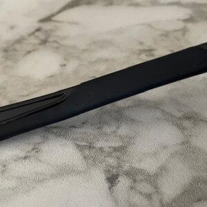 May include: Close-up of a black eyeglass arm. The arm is a sleek, rectangular shape with a slight curve. The surface appears smooth and matte, with a small, subtle design element near the hinge. The background is a light, marbled surface.