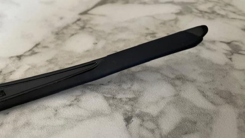 May include: Close-up of a black eyeglass arm. The arm is a sleek, rectangular shape with a slight curve. The surface appears smooth and matte, with a small, subtle design element near the hinge. The background is a light, marbled surface.