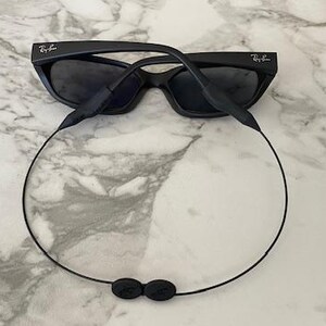 May include: Black Ray-Ban sunglasses featuring dark tinted lenses and a matte black frame. A thin black cord is attached to the temple arms, forming a loop that hangs below the frames, featuring a small black adjustment toggle at the bottom center.