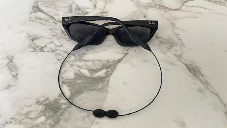 May include: Black Ray-Ban sunglasses featuring dark tinted lenses and a matte black frame. A thin black cord is attached to the temple arms, forming a loop that hangs below the frames, featuring a small black adjustment toggle at the bottom center.