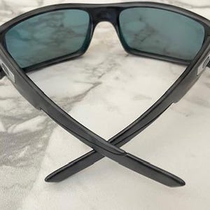 Puede incluir: Gafas de sol negras con lentes azules. Las gafas tienen una montura rectangular y un dise&ntilde;o moderno. Las patillas de las gafas est&aacute;n cruzadas, mostrando su forma. Las lentes parecen reflectantes.