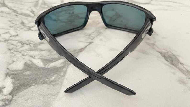 Puede incluir: Gafas de sol negras con lentes azules. Las gafas tienen una montura rectangular y un dise&ntilde;o moderno. Las patillas de las gafas est&aacute;n cruzadas, mostrando su forma. Las lentes parecen reflectantes.