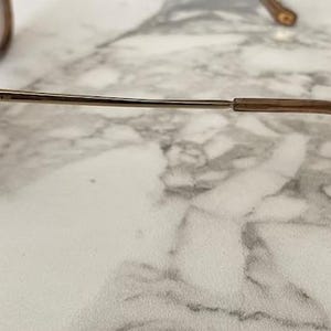 May include: Gold-tone metal sunglasses with brown lenses. The frame has a double bridge and a thin, straight arm that curves at the ear. The Coach logo is visible on the arm. The sunglasses are resting on a marble surface.