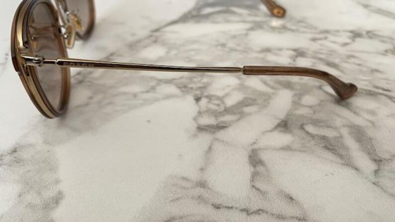 May include: Gold-tone metal sunglasses with brown lenses. The frame has a double bridge and a thin, straight arm that curves at the ear. The Coach logo is visible on the arm. The sunglasses are resting on a marble surface.