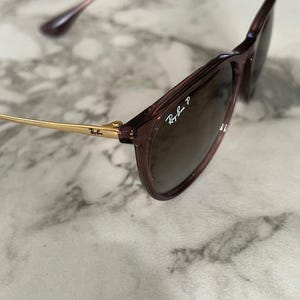 May include: Close-up of Ray-Ban sunglasses with a dark brown frame and lenses. The temples are gold-colored, and the Ray-Ban logo is visible on the lens. The sunglasses are angled, showing the details of the frame and lenses.