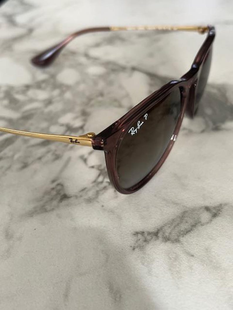 May include: Close-up of Ray-Ban sunglasses with a dark brown frame and lenses. The temples are gold-colored, and the Ray-Ban logo is visible on the lens. The sunglasses are angled, showing the details of the frame and lenses.