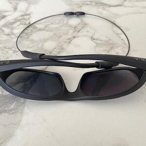 May include: Matte black sunglasses with dark tinted lenses and a thin, flexible cable strap attached to the temples. The frames feature a wrap-around design with the Ray-Ban logo visible on the side of the temple arms, shown in a folded position.