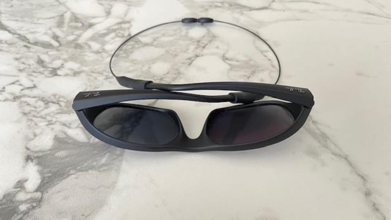 May include: Matte black sunglasses with dark tinted lenses and a thin, flexible cable strap attached to the temples. The frames feature a wrap-around design with the Ray-Ban logo visible on the side of the temple arms, shown in a folded position.