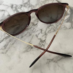 May include: Sunglasses with a dark brown frame and gradient lenses. The arms are gold and brown, with a sleek, modern design. The lenses have a brown tint, and the glasses are set against a marble background.