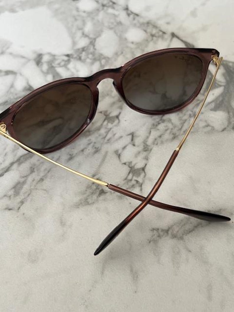 May include: Sunglasses with a dark brown frame and gradient lenses. The arms are gold and brown, with a sleek, modern design. The lenses have a brown tint, and the glasses are set against a marble background.