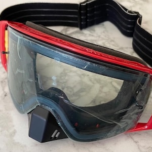 Può includere: Occhiali protettivi con montatura rossa, lente trasparente con pellicola protettiva e cinturino elastico nero. Gli occhiali includono un paranaso nero al centro, dettagli laterali gialli e un'imbottitura in schiuma di colore nero.