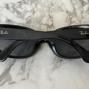 K&ouml;nnte beinhalten: Schwarze Sonnenbrille mit dem Ray-Ban-Logo auf den B&uuml;geln. Die Sonnenbrille hat einen dicken, rechteckigen Rahmen und dunkle Gl&auml;ser. Die Sonnenbrille liegt auf einer hellen, strukturierten Oberfl&auml;che.