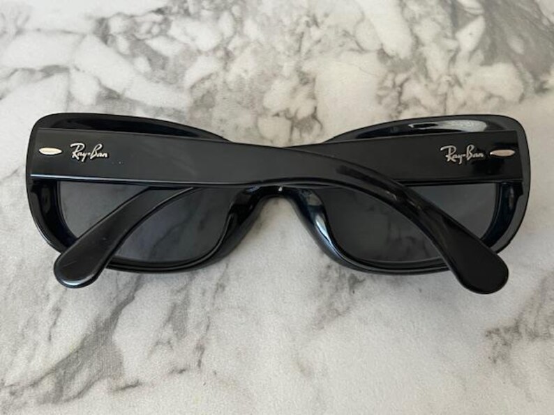 K&ouml;nnte beinhalten: Schwarze Sonnenbrille mit dem Ray-Ban-Logo auf den B&uuml;geln. Die Sonnenbrille hat einen dicken, rechteckigen Rahmen und dunkle Gl&auml;ser. Die Sonnenbrille liegt auf einer hellen, strukturierten Oberfl&auml;che.