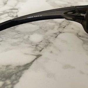 May include: Close-up of a pair of dark gray sunglasses. The sunglasses have a rectangular frame and dark lenses. The arm of the sunglasses has the text "OAKLEY" and other numbers printed on it. The sunglasses are on a white and gray marble surface.