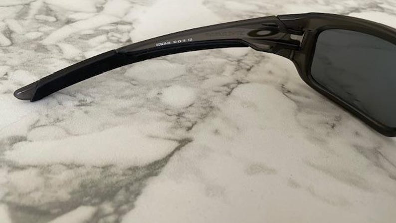 May include: Close-up of a pair of dark gray sunglasses. The sunglasses have a rectangular frame and dark lenses. The arm of the sunglasses has the text "OAKLEY" and other numbers printed on it. The sunglasses are on a white and gray marble surface.