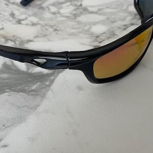 May include: Black sunglasses with orange-tinted lenses. The sunglasses have a sleek, sporty design with a glossy black frame. The lenses are a gradient color, transitioning from orange to a lighter hue. The sunglasses are shown on a marble surface.