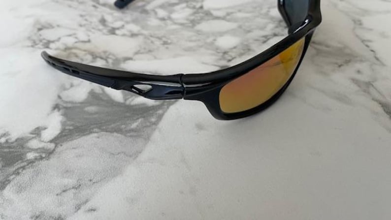 May include: Black sunglasses with orange-tinted lenses. The sunglasses have a sleek, sporty design with a glossy black frame. The lenses are a gradient color, transitioning from orange to a lighter hue. The sunglasses are shown on a marble surface.