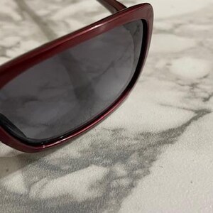 May include: Close-up of burgundy sunglasses with dark lenses. The sunglasses have a sleek, modern design with a slightly curved frame. The lenses are a dark gray color, providing shade and protection from the sun.