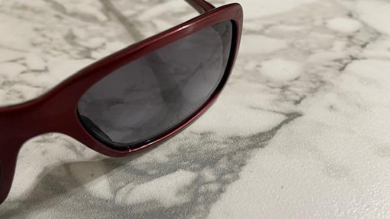 May include: Close-up of burgundy sunglasses with dark lenses. The sunglasses have a sleek, modern design with a slightly curved frame. The lenses are a dark gray color, providing shade and protection from the sun.