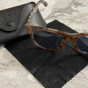 Puede incluir: Gafas de sol con montura marrón carey y lentes oscuras, sobre un paño de microfibra negro y junto a un estuche de cuero negro. El nombre de la marca "DIFF" está impreso en el estuche y el paño.
