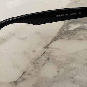 K&ouml;nnte beinhalten: Schwarze Sonnenbrille mit klassischem Design. Der Rahmen ist schwarz und die Gl&auml;ser sind dunkel. Der Brillenb&uuml;gel tr&auml;gt die Aufschrift "RB 2143 901 50022 3N". Die Brille liegt auf einer Marmoroberfl&auml;che.