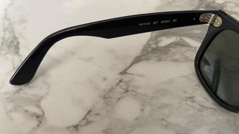 K&ouml;nnte beinhalten: Schwarze Sonnenbrille mit klassischem Design. Der Rahmen ist schwarz und die Gl&auml;ser sind dunkel. Der Brillenb&uuml;gel tr&auml;gt die Aufschrift "RB 2143 901 50022 3N". Die Brille liegt auf einer Marmoroberfl&auml;che.