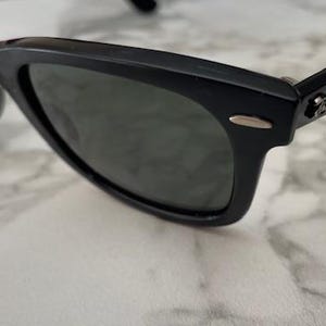 K&ouml;nnte beinhalten: Schwarze Sonnenbrille mit dunklen Gl&auml;sern. Die Sonnenbrille hat ein klassisches, rechteckiges Rahmendesign. Das Ray-Ban-Logo ist auf dem B&uuml;gel sichtbar. Die Sonnenbrille liegt auf einer wei&szlig;-grauen Marmoroberfl&auml;che.