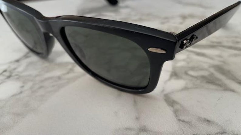 K&ouml;nnte beinhalten: Schwarze Sonnenbrille mit dunklen Gl&auml;sern. Die Sonnenbrille hat ein klassisches, rechteckiges Rahmendesign. Das Ray-Ban-Logo ist auf dem B&uuml;gel sichtbar. Die Sonnenbrille liegt auf einer wei&szlig;-grauen Marmoroberfl&auml;che.