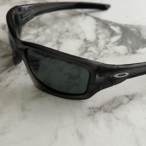 May include: Dark grey sunglasses with a wraparound design. The lenses are a dark grey colour, and the frames have a subtle, translucent appearance. The Oakley logo is visible on the temple.