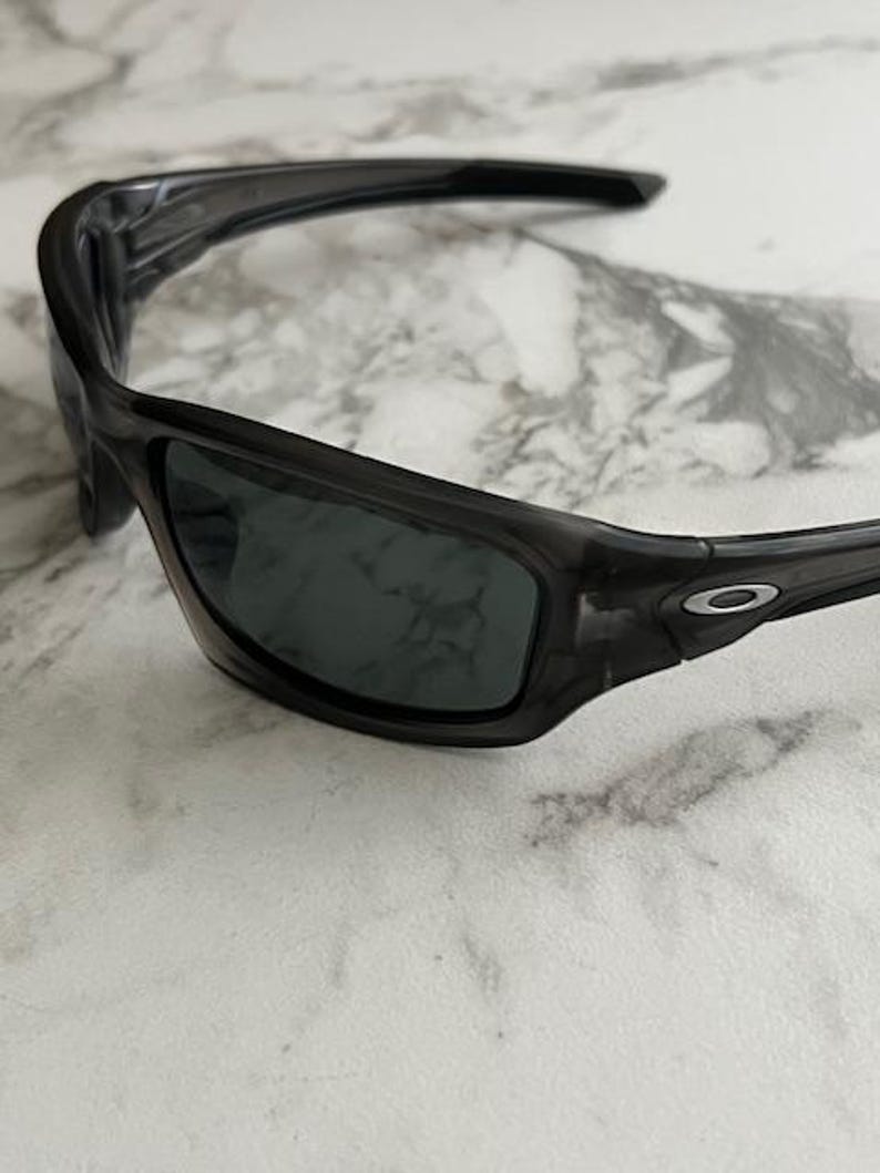 May include: Dark grey sunglasses with a wraparound design. The lenses are a dark grey colour, and the frames have a subtle, translucent appearance. The Oakley logo is visible on the temple.