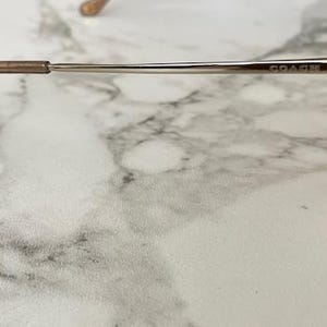 May include: Close-up of Coach eyeglasses. The glasses have a brown and gold color scheme. The frame is thin and metallic, with the brand name "COACH" visible on the temple. The earpiece is a darker brown.