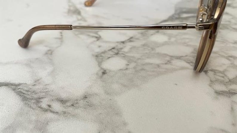 May include: Close-up of Coach eyeglasses. The glasses have a brown and gold color scheme. The frame is thin and metallic, with the brand name "COACH" visible on the temple. The earpiece is a darker brown.