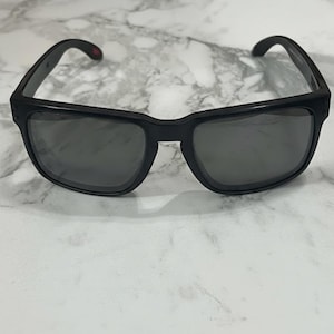 May include: Black sunglasses with a rectangular frame and dark lenses. The sunglasses have a small red accent on the temple. The frame is made of a matte black material.