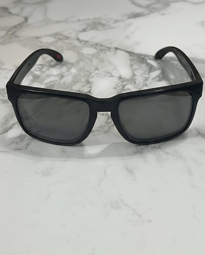 May include: Black sunglasses with a rectangular frame and dark lenses. The sunglasses have a small red accent on the temple. The frame is made of a matte black material.