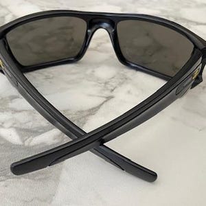 May include: Black sunglasses with dark lenses. The sunglasses have a rectangular frame and are positioned with the arms crossed. The lenses are a dark grey colour, and the frame is a matte black. The sunglasses are on a white marble surface.